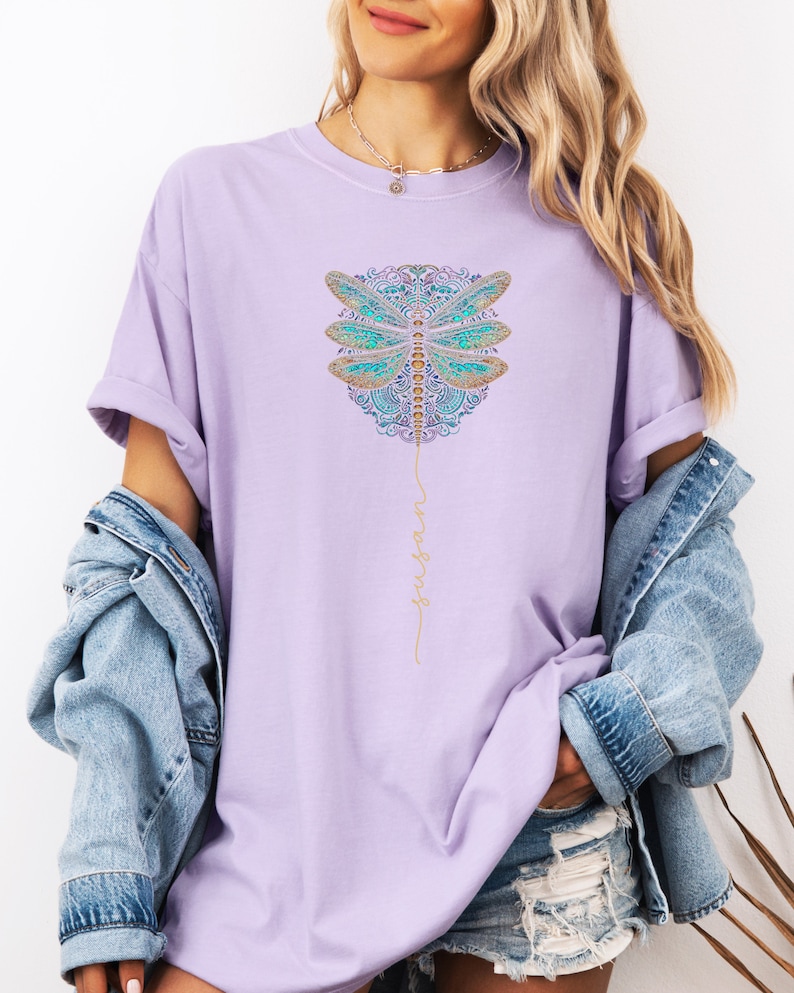 May include: A lavender t-shirt featuring a gold and blue dragonfly graphic with the word "trust" written vertically in gold script.