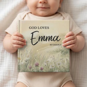 May include: A personalized children's book with the title "God Loves Emma So Deeply." The cover features a watercolor illustration of a meadow with wildflowers and tall grass. The book is held by a baby.