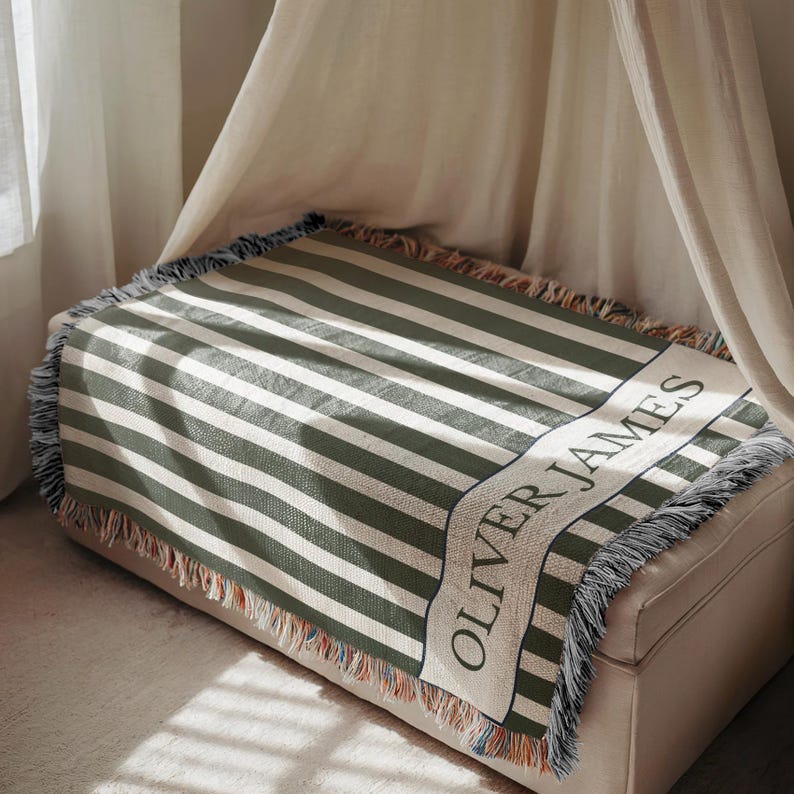 May include: A woven blanket with olive green and white stripes, fringed edges, and the text "OLIVER JAMES" in a banner. The blanket is draped over a beige ottoman, with a canopy in the background.
