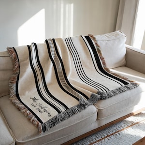 May include: Cream-colored throw blanket with black vertical stripes and fringed edges. The blanket is draped over a beige sofa. The text "The Johnsons EST. 2025" is visible in the corner.