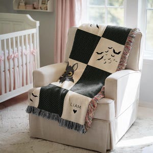 May include: A black and cream-colored baby blanket with a patchwork design featuring a cartoon dragon, bats, and polka dots. The blanket is draped over a white armchair in a nursery setting. The name "LIAM" and a heart are printed on the blanket.