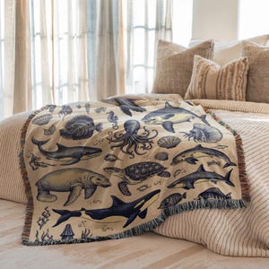 Puede incluir: Una manta beige con una ilustraci&oacute;n azul marino de varias criaturas marinas, incluyendo tiburones, un pulpo y una tortuga. La manta tiene un borde con flecos y est&aacute; colocada sobre una cama con almohadas de tonos neutros.