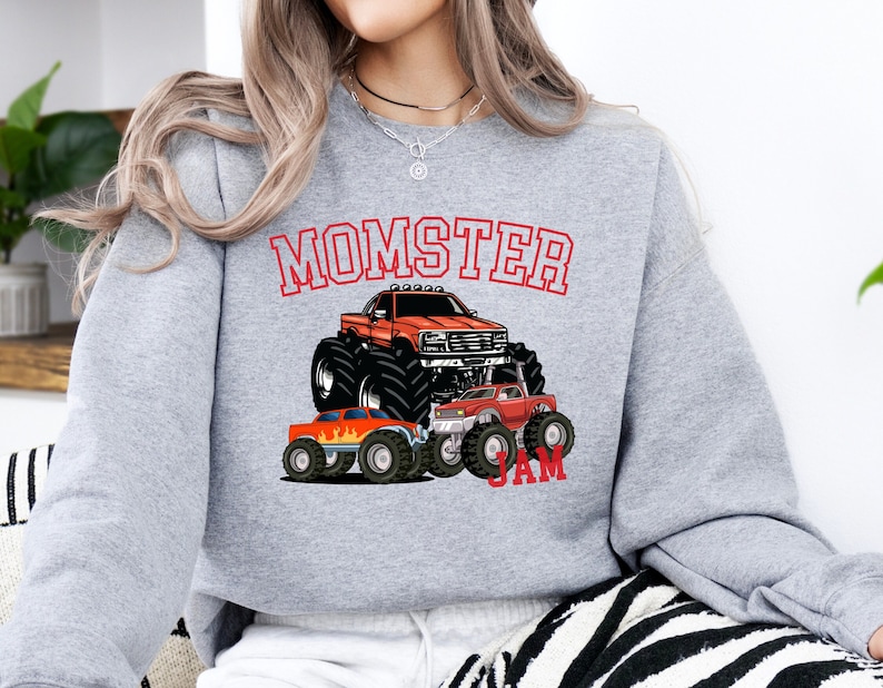 May include: Gray sweatshirt with the text "MOMSTER JAM" in red. The sweatshirt features a cartoon illustration of three monster trucks, one large and two smaller, with flames.