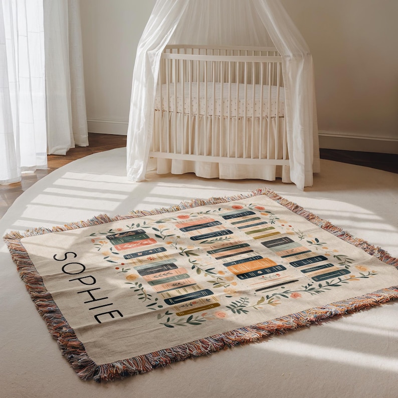May include: A cream-colored woven throw blanket with a colorful fringe border. The blanket features a pattern of stacked books with floral accents and the name "Sophie" in black lettering.
