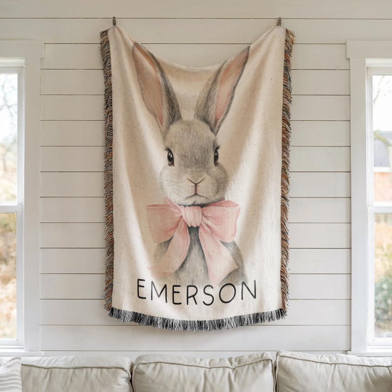 Puede incluir: Una manta tejida beige con una ilustraci&oacute;n detallada de un conejo gris con una pajarita rosa. El nombre "EMERSON" est&aacute; impreso en negro en la parte inferior. La manta tiene un borde con flecos y est&aacute; colgada en una pared blanca.