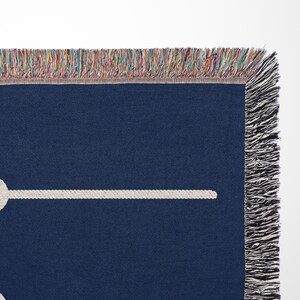 May include: A dark navy blue woven throw blanket featuring a white rope graphic tied in a knot. The blanket has a decorative fringe edge with multicoloured threads woven into the top border and solid black fringe along the side edge.