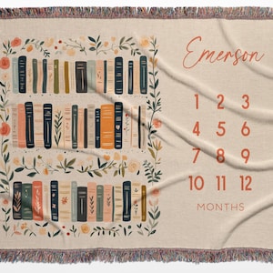 Op de afbeelding: Beige geweven sprei met een boekenplankontwerp en bloemaccenten. De sprei heeft een boekenplankillustratie met verschillende kleuren boeken en de naam "Emerson" in een scriptlettertype. Cijfers 1-12 worden weergegeven voor maandelijkse foto's.