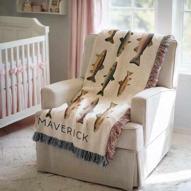 May include: A cream-colored throw blanket with a fish pattern and the name "MAVERICK" in black lettering. The blanket is draped over a white armchair. The blanket has a fringed edge and is in a nursery setting.