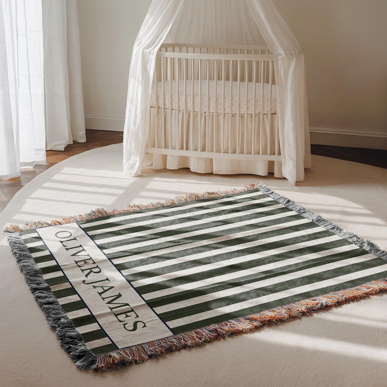 May include: A woven blanket with olive green and white stripes, fringed edges, and the name "OLIVER JAMES" in a white rectangular panel. The blanket is on a light-colored rug in a nursery setting.