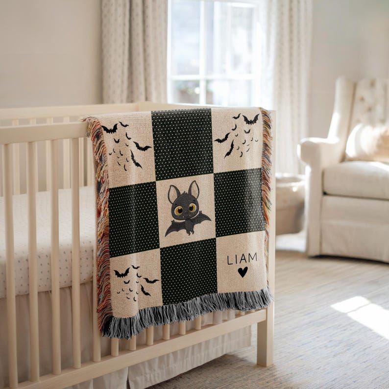 May include: A beige and black checkered baby blanket with a bat theme. The blanket features a cute bat illustration in the center square, bat silhouettes, and the name "LIAM" with a heart. The blanket is draped over a white crib.