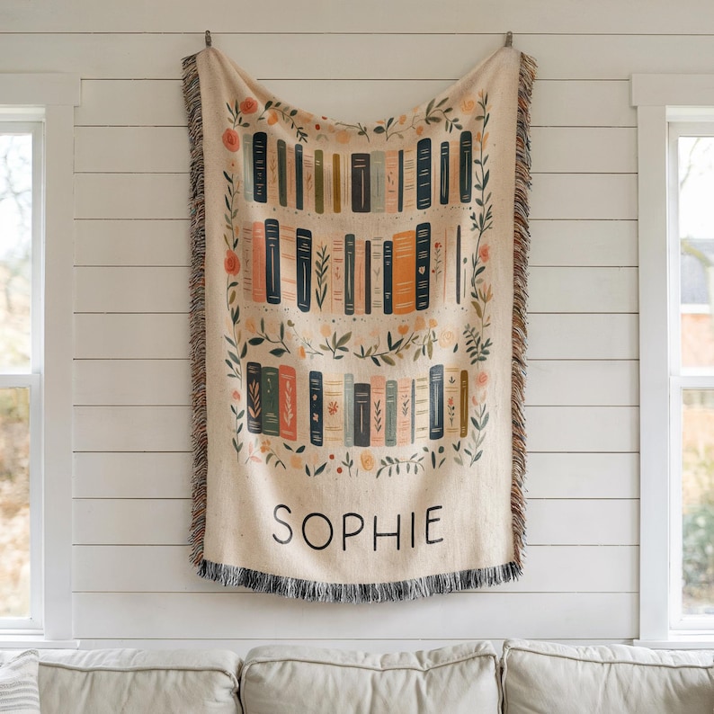 May include: A cream-colored woven throw blanket with a colorful illustration of books on a bookshelf. The blanket has a fringe border and the name "SOPHIE" is printed in black at the bottom.