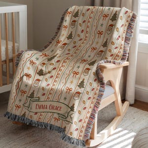 May include: A woven throw blanket featuring a cream background with vertical stripes of Christmas trees, Santa Claus faces, bows, and string lights. A decorative banner at the bottom displays the name Emma Grace in red lettering with fringed edges.
