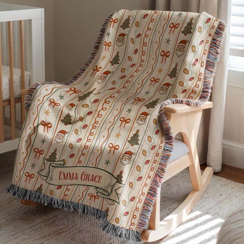 May include: A woven throw blanket featuring a cream background with vertical stripes of Christmas trees, Santa Claus faces, bows, and string lights. A decorative banner at the bottom displays the name Emma Grace in red lettering with fringed edges.