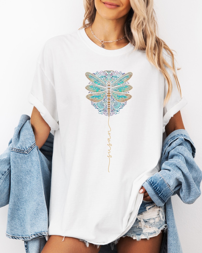 May include: White t-shirt featuring a blue and gold dragonfly design with the words "fly high" written in gold script.