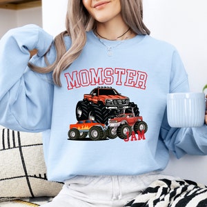 May include: Light blue sweatshirt with a red, white, and blue monster truck graphic. The text "MOMSTER JAM" is printed on the sweatshirt.