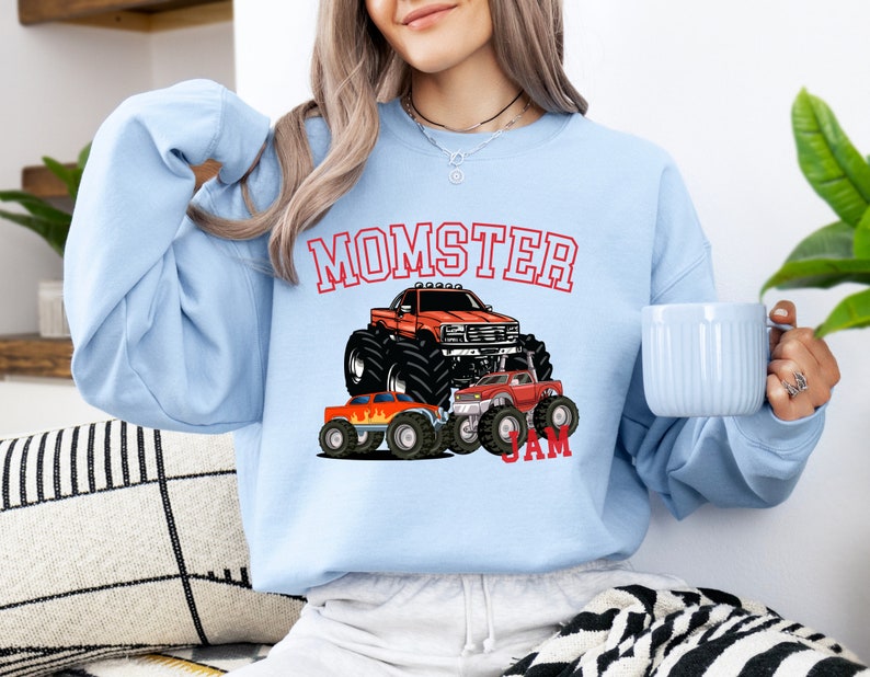 May include: Light blue sweatshirt with a red, white, and blue monster truck graphic. The text "MOMSTER JAM" is printed on the sweatshirt.