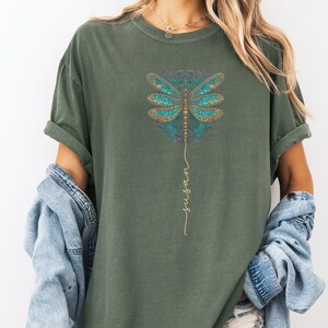 May include: A green t-shirt featuring a blue and gold dragonfly design with the word "trust" written vertically beneath it.