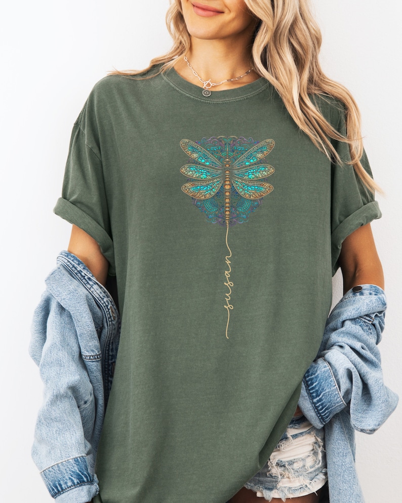 May include: A green t-shirt featuring a blue and gold dragonfly design with the word "trust" written vertically beneath it.
