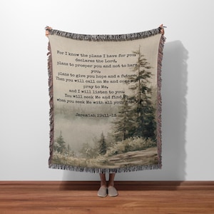 May include: A woven throw blanket featuring a forest scene with text from Jeremiah 29:11-13. The blanket has a fringed edge and a neutral color palette with shades of beige, brown, and green. The text is in a dark font.