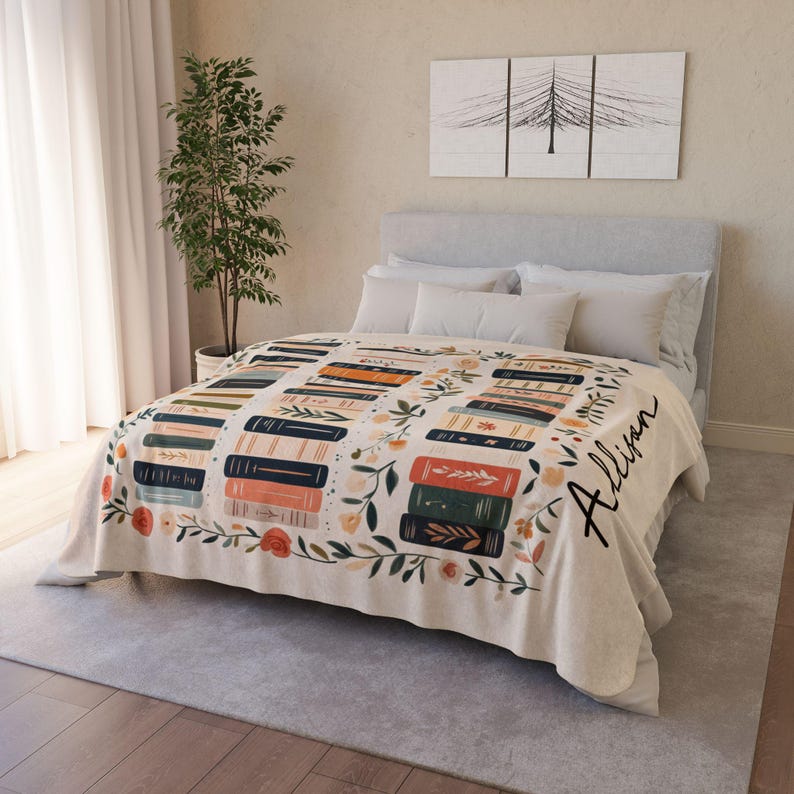 May include: A cream-colored fleece blanket with a personalized name "Allison" in black script. The blanket features a pattern of stacked books with floral accents in shades of orange, green, blue, and pink.