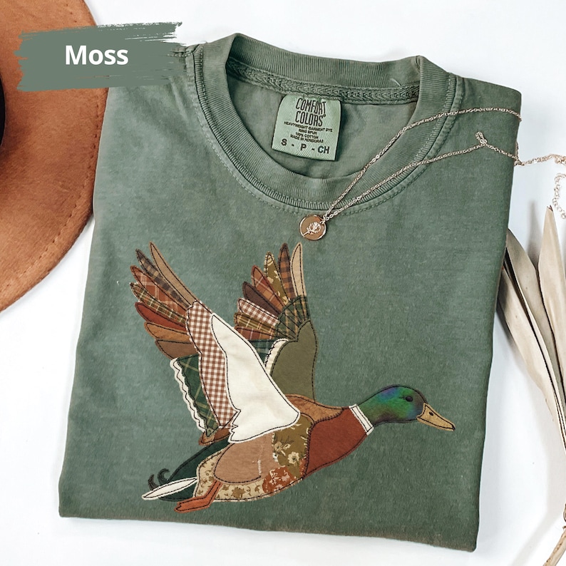 K&ouml;nnte beinhalten: Ein moosgr&uuml;nes T-Shirt mit einem aufgen&auml;hten Entenmotiv. Die Fl&uuml;gel der Ente bestehen aus verschiedenen gemusterten Stoffen. Das T-Shirt hat einen Rundhalsausschnitt und kurze &Auml;rmel. Eine silberne Halskette mit Anh&auml;nger liegt auf dem T-Shirt.