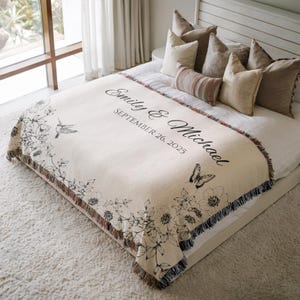 May include: Cream-colored woven throw blanket with black floral and hummingbird design. Features the names "Emily & Michael" and the date "September 26, 2025" in elegant script. Edged with a fringed border.