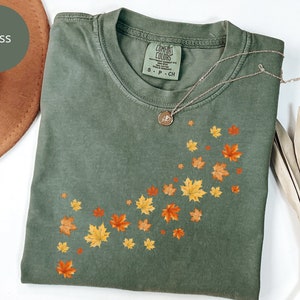 May include: Moss green t-shirt with a crew neck, featuring a design of falling autumn leaves in shades of yellow, orange, and red. A gold necklace with a pendant rests on the shirt. The shirt is folded and placed on a white surface.