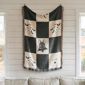 May include: A black and cream-colored throw blanket with a bat theme. The blanket features a checkerboard pattern with black polka dots and bat silhouettes. A central square showcases a cute bat illustration with the name "LIAM" and a heart symbol.