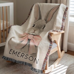Puede incluir: Una manta tejida con una ilustraci&oacute;n de un conejo gris y un lazo rosa. El nombre "EMERSON" est&aacute; impreso en negro. La manta tiene un borde con flecos y est&aacute; sobre una mecedora.