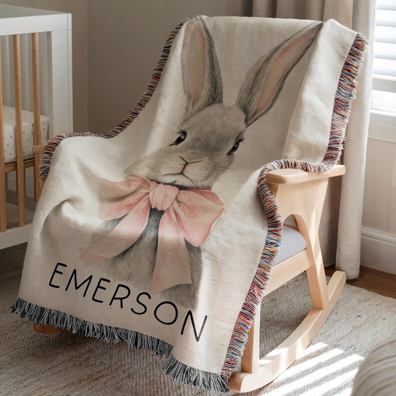 Puede incluir: Una manta tejida con una ilustraci&oacute;n de un conejo gris y un lazo rosa. El nombre "EMERSON" est&aacute; impreso en negro. La manta tiene un borde con flecos y est&aacute; sobre una mecedora.