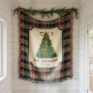 May include: A woven throw blanket featuring a red, black, and green plaid pattern with a central cream-colored panel. The panel displays a green Christmas tree with a red bow, the text "The Johnsons" and "Est. 2026," and has fringed edges.