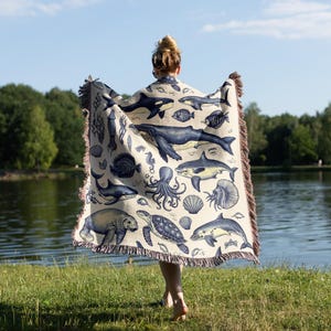 Puede incluir: Una manta beige con un dise&ntilde;o n&aacute;utico azul y blanco que representa diversas criaturas marinas, como ballenas, tiburones y pulpos. La manta tiene un borde con flecos y se muestra frente a un lago y &aacute;rboles.