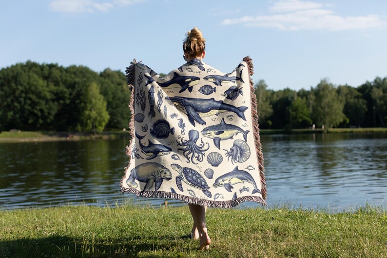 Puede incluir: Una manta beige con un dise&ntilde;o n&aacute;utico azul y blanco que representa diversas criaturas marinas, como ballenas, tiburones y pulpos. La manta tiene un borde con flecos y se muestra frente a un lago y &aacute;rboles.