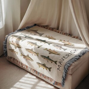 May include: A cream-colored throw blanket featuring a repeating pattern of various fish illustrations in shades of brown, green, and gray. The word "MAVERICK" is printed in black on the lower right corner. The blanket has a fringed edge.