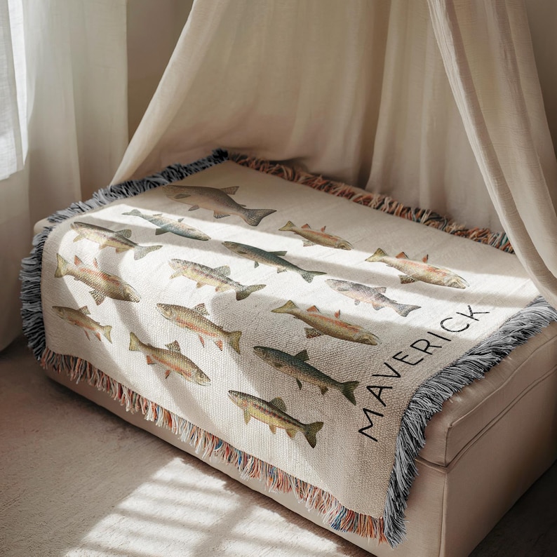 May include: A cream-colored throw blanket featuring a repeating pattern of various fish illustrations in shades of brown, green, and gray. The word "MAVERICK" is printed in black on the lower right corner. The blanket has a fringed edge.