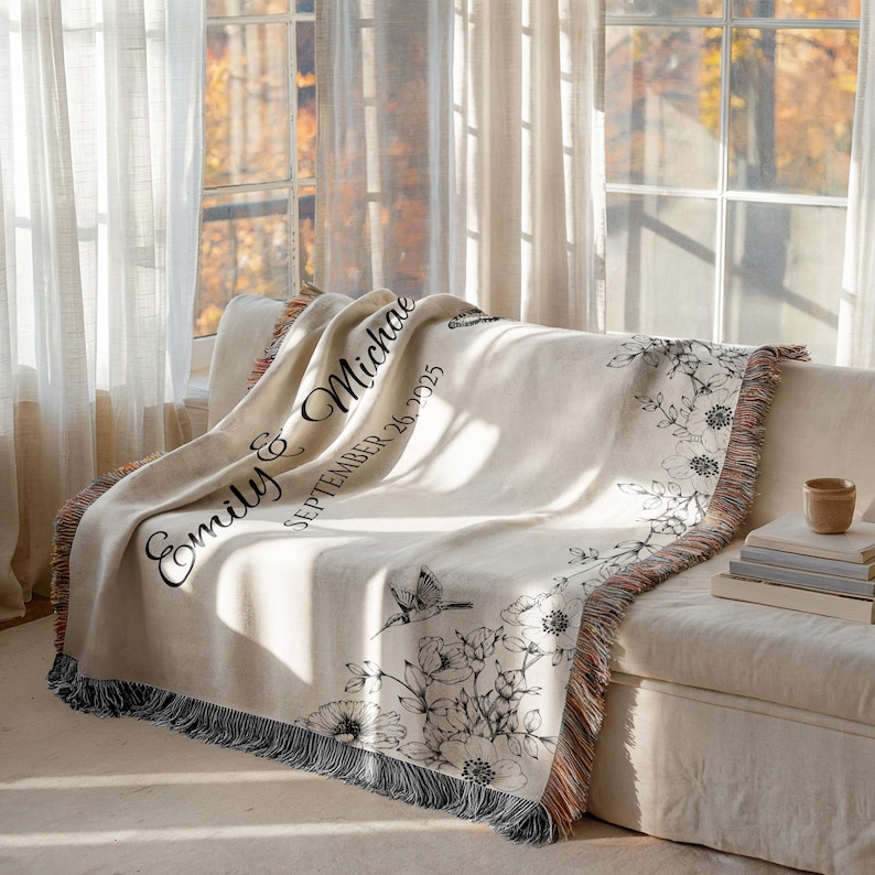 May include: Cream-colored woven throw blanket draped over a sofa. The blanket features black floral and hummingbird designs, with the names "Emily & Michael" and the date "September 26, 2025" printed on it. The blanket has a fringed edge.