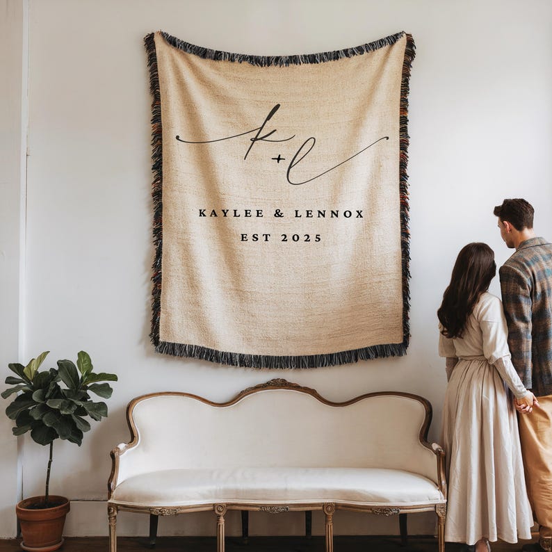 Op de afbeelding: Een beige geweven sprei met zwarte franjes, gepersonaliseerd met de namen "Kaylee & Lennox" en het jaar "2025" in zwart schrift. De sprei hangt aan een witte muur boven een witte antieke bank.