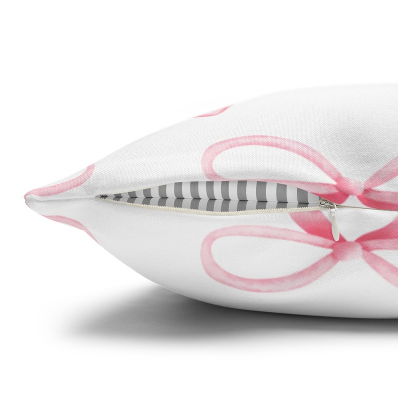 Puede incluir: Almohada blanca con un patr&oacute;n de lazo rosa en acuarela. La almohada tiene una cremallera y un forro a rayas gris y blanco.