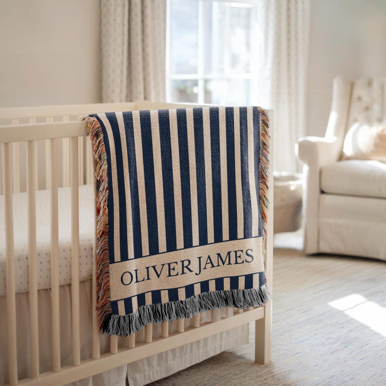 May include: A navy and cream striped baby blanket with the name "OLIVER JAMES" in black lettering. The blanket is draped over a white crib and has fringed edges. The nursery setting includes a white armchair and a window.