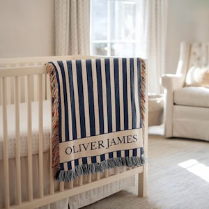 Op de afbeelding: Een babydeken met marineblauwe en cr&egrave;mekleurige strepen met de naam "OLIVER JAMES" in zwarte letters. De deken is over een witte wieg gedrapeerd en heeft franjes. De kinderkamerinrichting omvat een witte fauteuil en een raam.