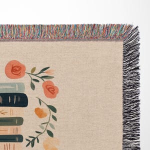 May include: A woven tapestry throw blanket with a cream background and a stack of books with floral accents. The blanket has a black fringe border.