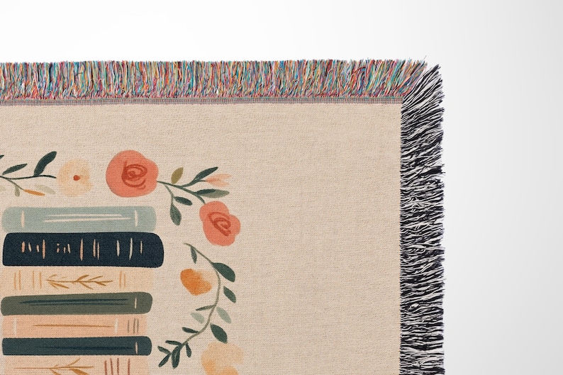 May include: A woven tapestry throw blanket with a cream background and a stack of books with floral accents. The blanket has a black fringe border.