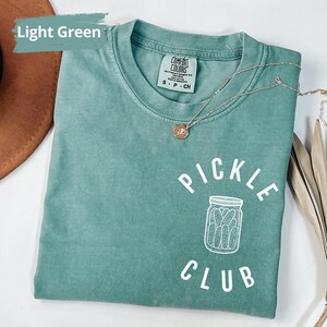 Peut inclure: T-shirt vert clair avec le texte "Pickle Club" en blanc. Le t-shirt pr&eacute;sente une illustration blanche d'un pot de cornichons.