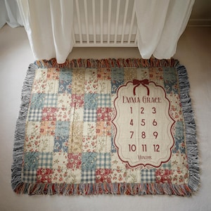 May include: A patchwork baby blanket with a floral and gingham pattern in shades of red, blue, and cream. The blanket has a decorative fringe and a label with the name "Emma Grace" and numbers 1-12 for monthly milestones.