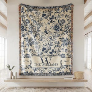 May include: A large, cream-colored throw blanket with a blue floral pattern. The blanket has a rectangular label with the text "W THE WILLIAMS ESTD 2026". The edges are fringed.