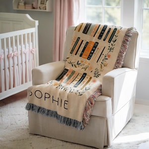May include: A cream-colored woven throw blanket with a colorful pattern of stacked books and floral accents. The blanket has fringe on all sides and the word "Sophie" is embroidered on the bottom corner.