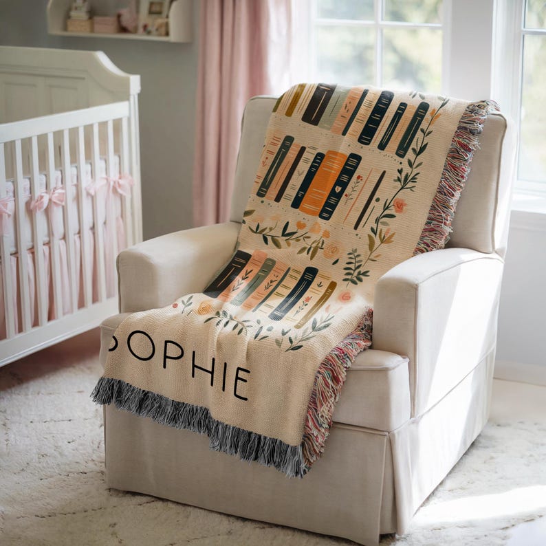 May include: A cream-colored woven throw blanket with a colorful pattern of stacked books and floral accents. The blanket has fringe on all sides and the word "Sophie" is embroidered on the bottom corner.