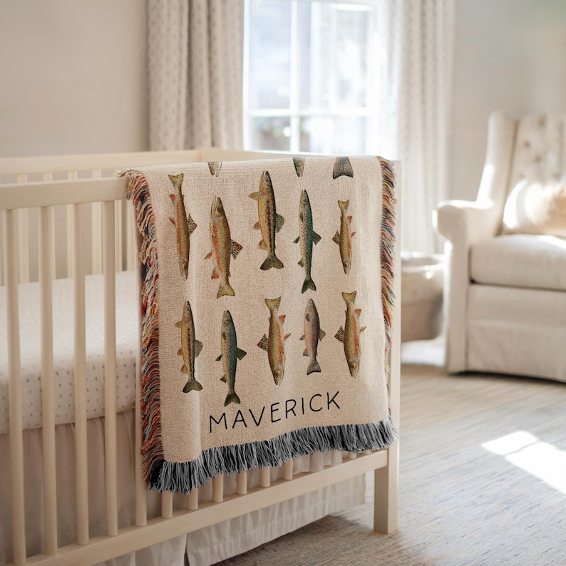 May include: A cream-colored baby blanket with a repeating pattern of colorful fish. The blanket is draped over a white crib and has a fringe along the bottom. The name "MAVERICK" is printed in black letters on the blanket.