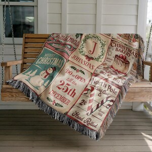 May include: A woven throw blanket featuring a grid of Christmas-themed illustrations including a snowman, a wreath with the letter J, a cake labeled Christmas Bakery, and candy canes with text reading Do not open till the 25th December.