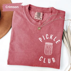 Peut inclure: Un t-shirt rouge cramoisi avec l'inscription "Pickle Club" en blanc. Le t-shirt pr&eacute;sente une illustration blanche d'un pot de cornichons.